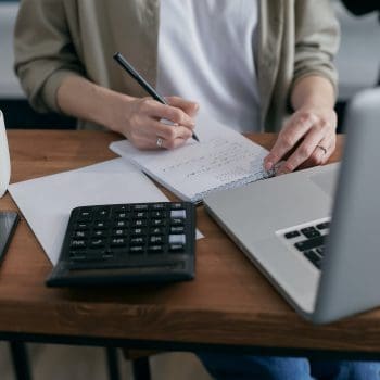 Person at a laptop writing on a notepad alongside a black calculator