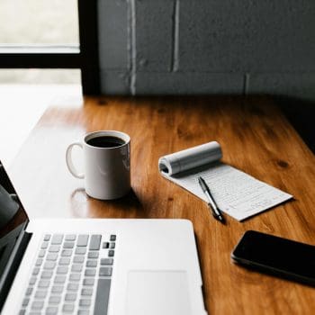 A desk with a laptop, coffee cup, cell phone, and notepad