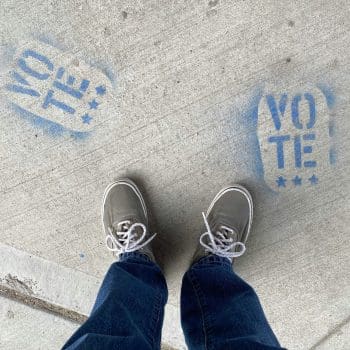 A person standing over two spray-painted logos that say "VOTE" on the sidewalk